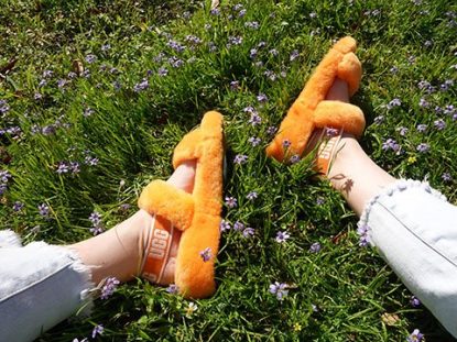 Feet shown wearing orange UGG Oh Yeah Slide Sandals, while resting in a patch of bright green grass and purple flowers.